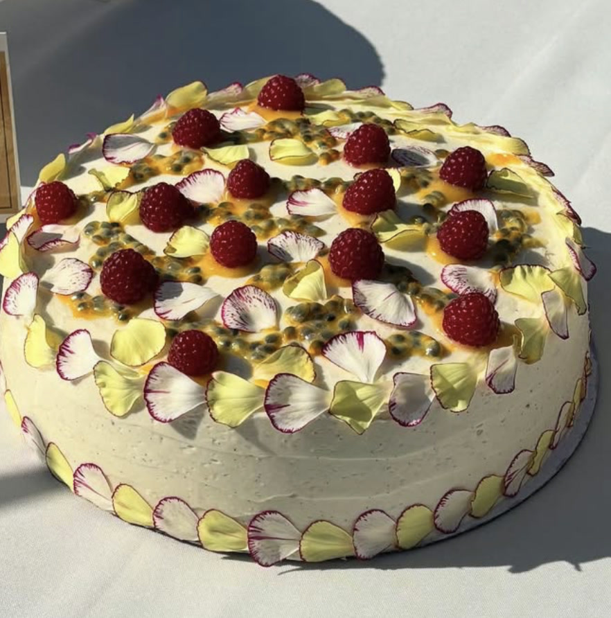 A round cake topped with raspberries, passionfruit, and yellow and purple edible petals